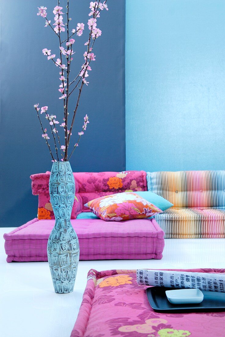 Combination of floor cushions in pastel shades against blue wall; artistic floor vase of Japanese cherry blossom in foreground