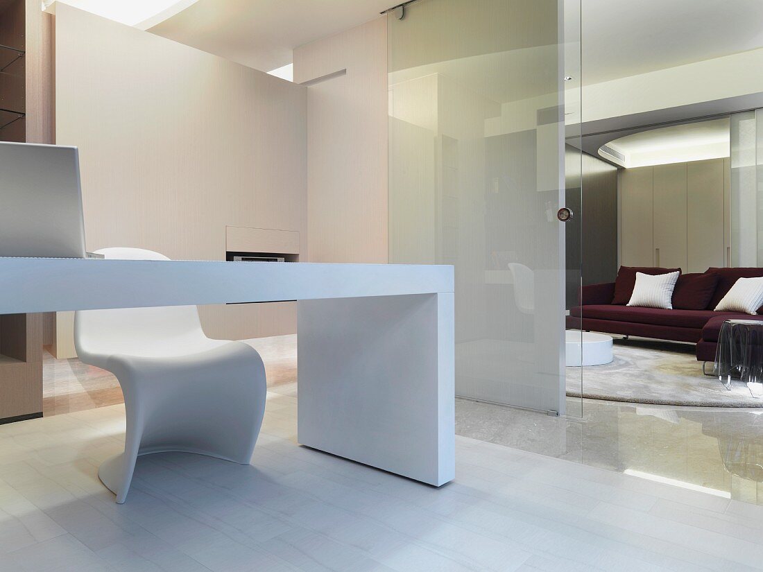 Weisser, minimalistischer Schreibtisch und Schalenstuhl aus weißem Kunststoff neben offener Glastür mit Blick auf Sofa