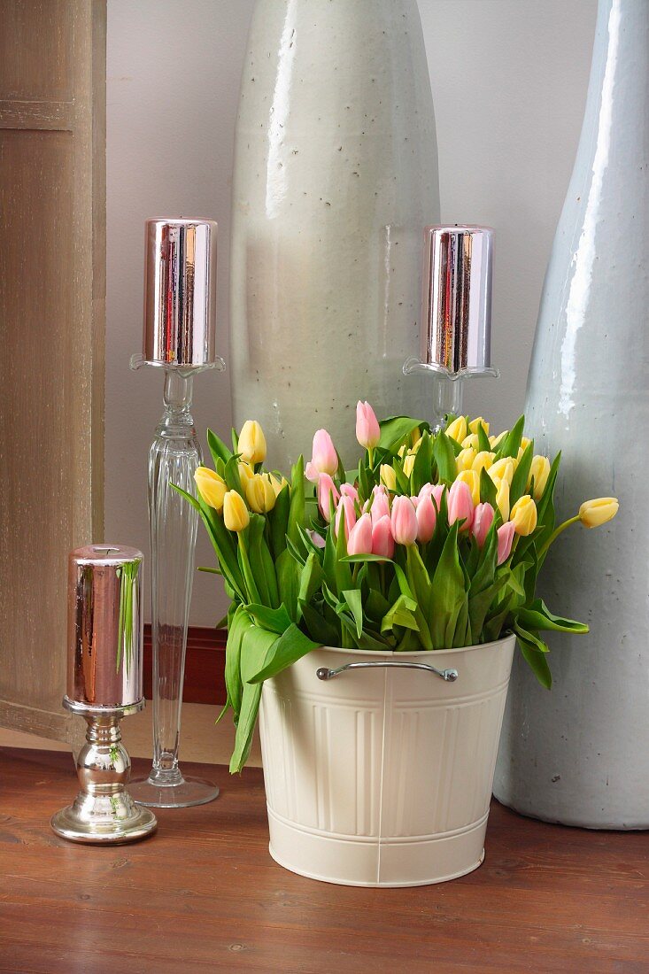 Gelbe und rosafarbene Tulpen in hellem Blecheimer dekoriert mit festlich glänzenden Kerzen auf verschiedenen Ständern