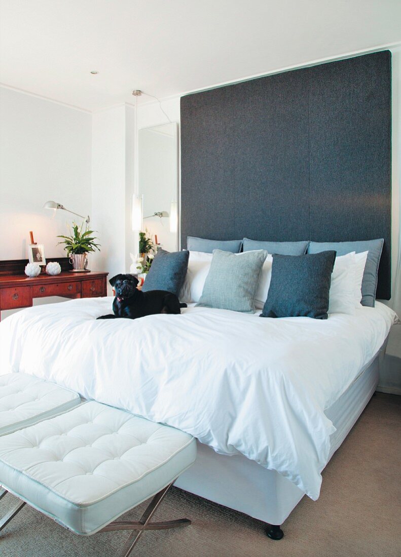 White double bed with tall headboard upholstered in dark grey and white, designer stools at foot