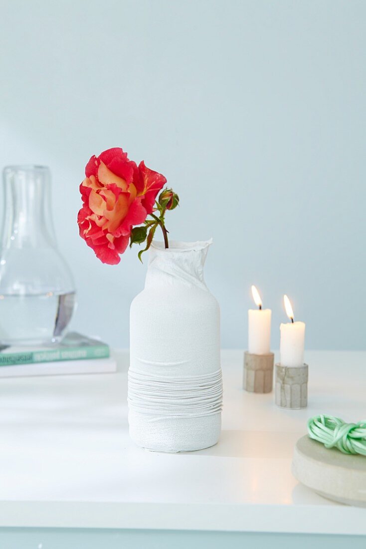 A porcelain vase covered with a stocking and painted white, decorated with a rose next to candles