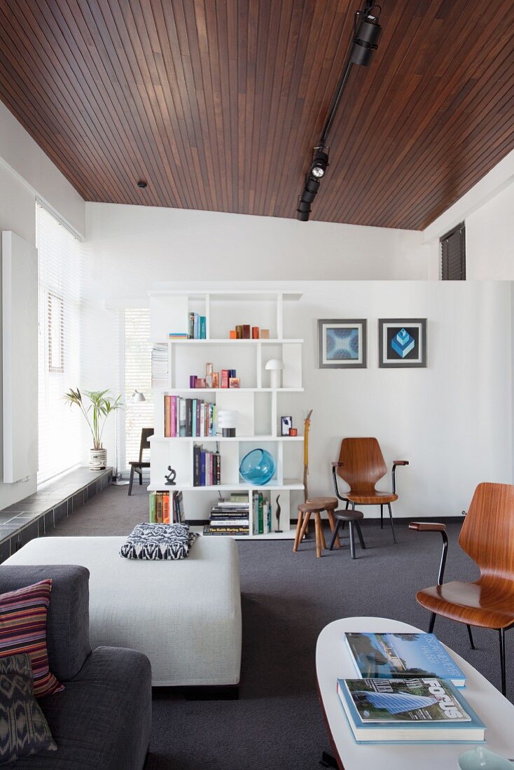 Wohnraum mit Holzdecke, im Vordergrund heller Polstertisch und Retro Stühle vor weißer Raumteilwand