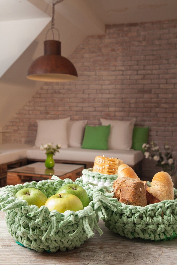Grüne Häkelkörbe für Obst und Brot auf Holztisch, im Hintergrund Sitzecke vor Backsteinwand