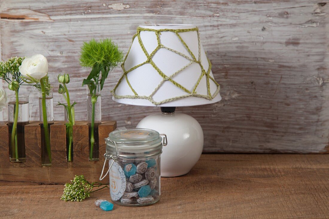 Green chain-stitch net cover on white lampshade next to single flowers in test tube vases