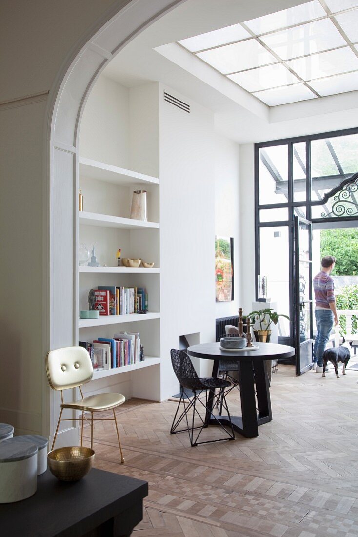 Round table, wire mesh chair, white fitted shelving in open-plan, high-ceilinged interior with traditional, elegant ambiance; man and dog in front of open terrace doors