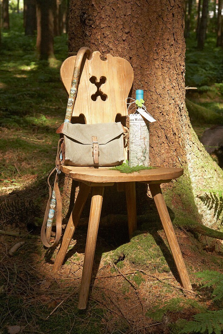 Wanderutensilien auf Holzstuhl mit geschnitzter Rückenlehne vor Baum im Wald, Weinflasche mit Landkarte umwickelt