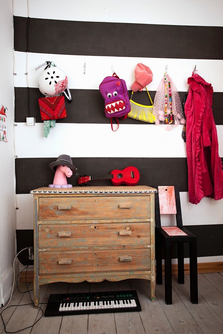 Vintage Kommode vor schwarz-weiss gesgtreifter Wand, bunte Kinderaccessoires an Garderobenhaken aufgehängt