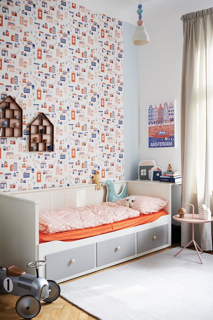Grau-weißes Holzbett mit Bettschubladen und orangefarbenem Spannbetttuch vor Motivtapete in Kinderzimmer