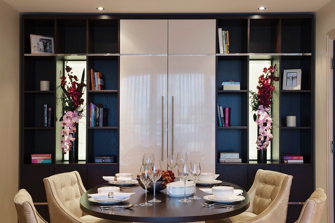 Schrankwand mit Regalflächen, geschlossenen Fronten und Spiegelnischen mit eleganten Blumensträussen; davor ein gedeckter Tisch mit Samtsesseln