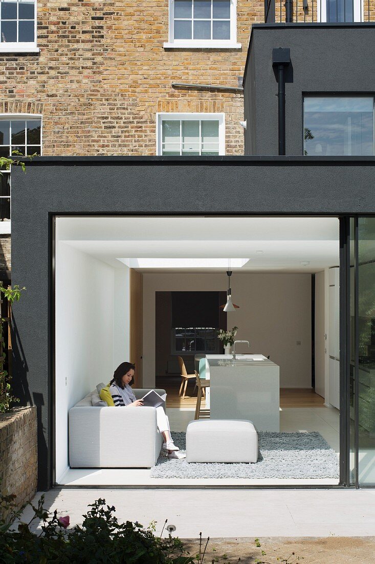 Modern, dark grey extension to Georgian terrace house with open, sliding glass wall leading to garden; woman reading on pale grey sofa
