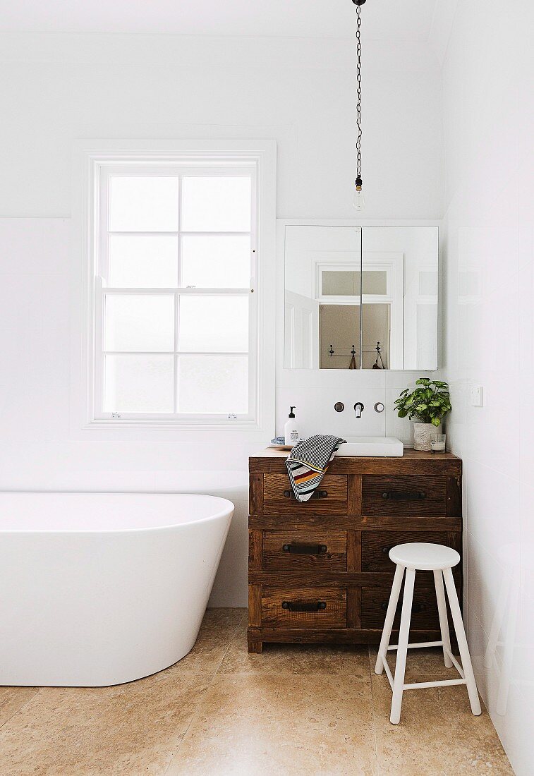 Vintage vanity furniture made of rustic wood in the corner of a bathroom next to a free-standing white bathtub