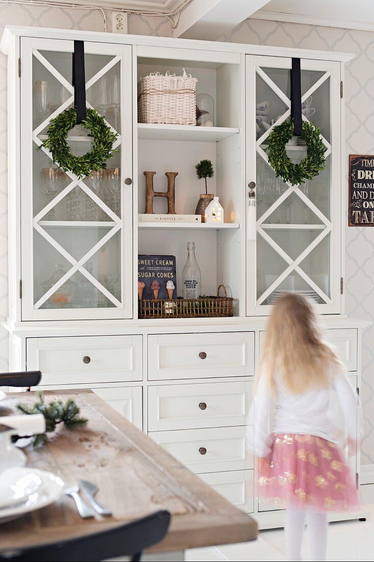 Girl walking past white kitchen dresser decorated with wreaths of box leaves