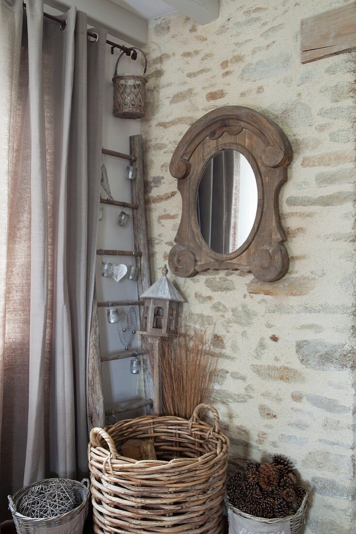 Weidenkorb mit Tannenzapfen und Holzscheiten in Zimmerecke mit Natursteinwand und rustikalem Spiegel