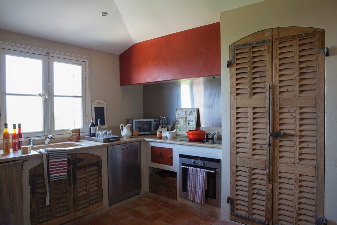 Rustic masonry kitchen counter below window and fitted cupboard with wooden louvre doors
