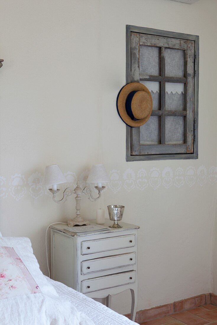 Antique bedside cabinet next to bed and straw hat hung on interior window in bedroom