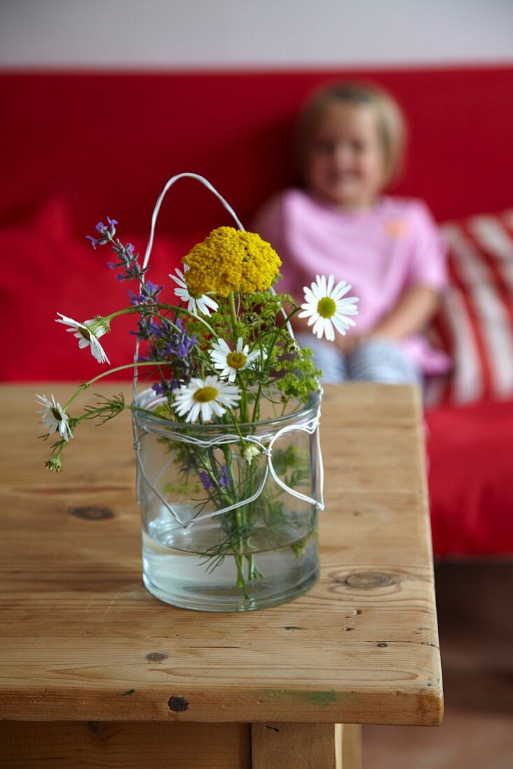 Wiesenblumenstrauss im Wasserglas auf Holztisch, dahinter Mädchen auf Sofa