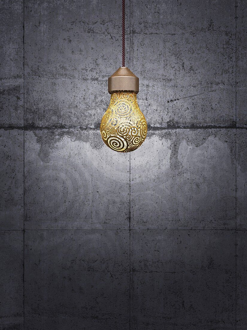 Light bulb with gold ornamentation hanging in front of grey concrete wall