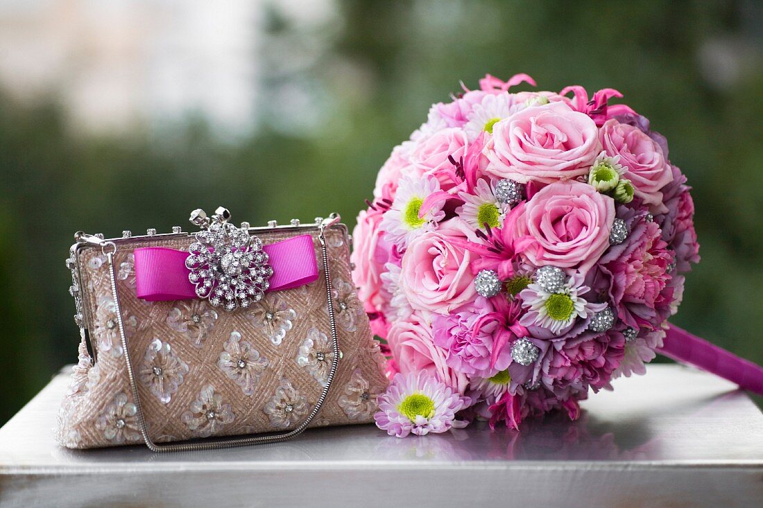 Romantischer pinkfarbener Hochzeitsstrauß neben elegantem Täschchen mit glitzernder Brosche auf Vintagekoffer