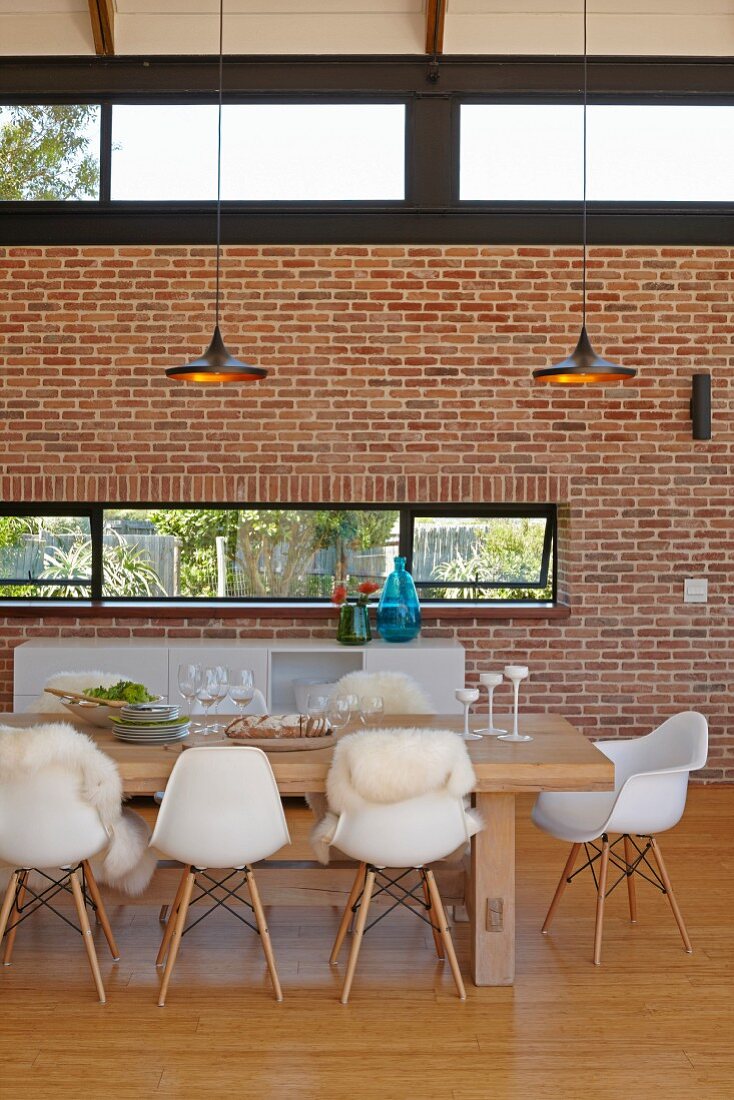 Essplatz mit Klassikerstühlen um Massivholztisch oberhalb Designerleuchten, gegenüber Ziegelwand mit Oberlichtern in modernem Wohnraum