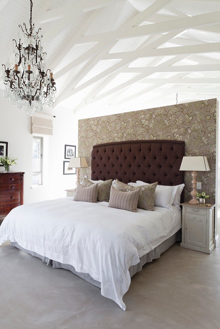 Heller Schlafbereich mit tapezierter Wandscheibe und dunkelbraun gepolstertem Bettkopfteil, Kronleuchter und weiß gestrichener Dachkonstruktion