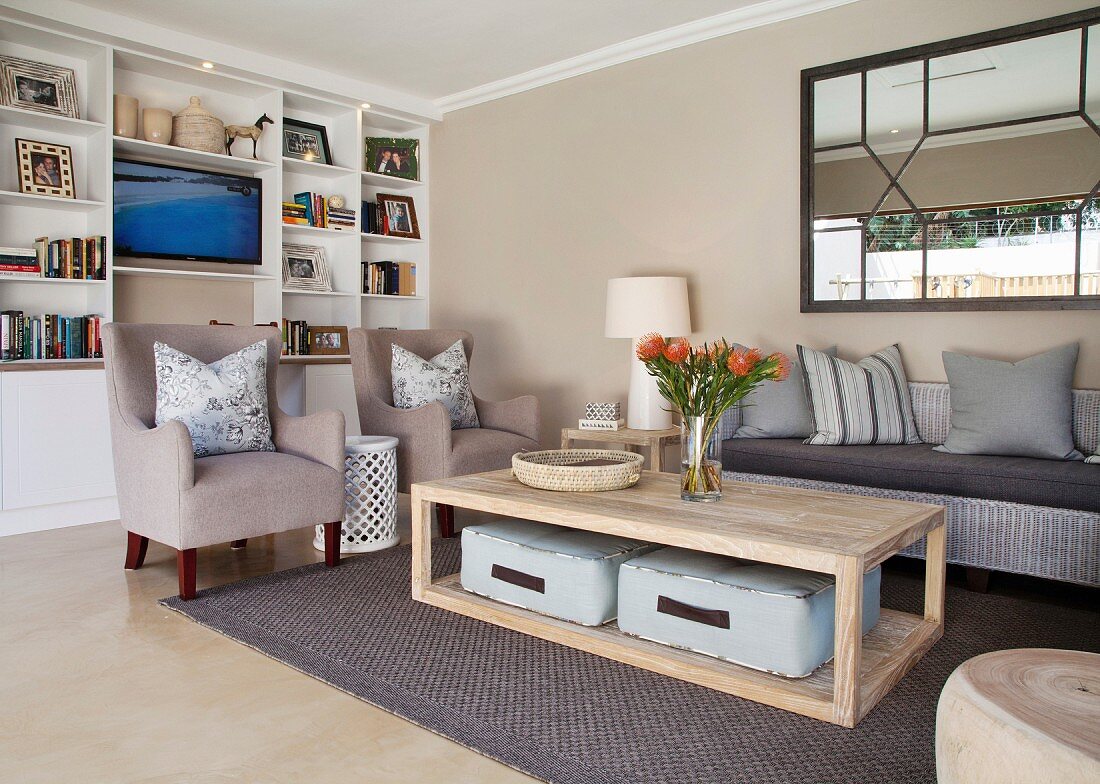 Gemütliches Wohnzimmer mit weißem Einbauregal, beigefarbenen Sesseln, Rattancouch und Bodenkissen unter Couchtisch