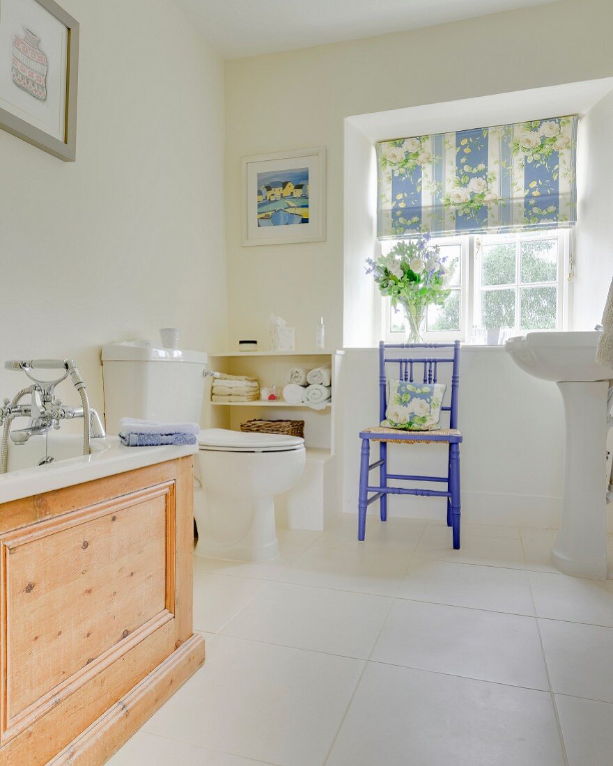 Ländliches Bad mit weißem Fliesenboden, Badewanne mit Holzverkleidung und Stuhl vor Sprossenfenster