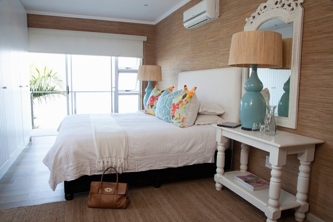 Schlafzimmer mit weißem Einbauschrank, türkisfarbenen Tischleuchten und mit Kissen dekoriertes Bett vor tapezierter Wand