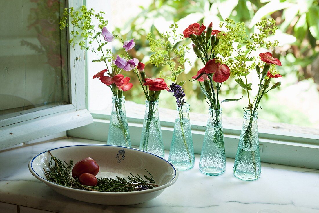 Rosemary sprig in bowl and flowers arranged in small bottles on windowsill
