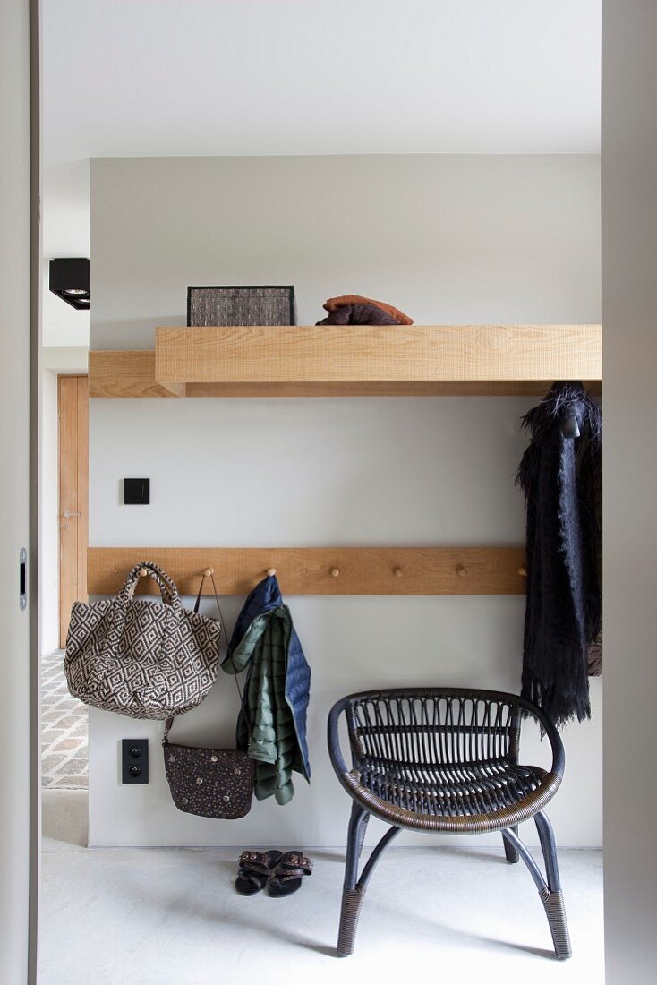 Wicker chair below minimalist cloakroom rack with bags and clothes hanging from coat pegs