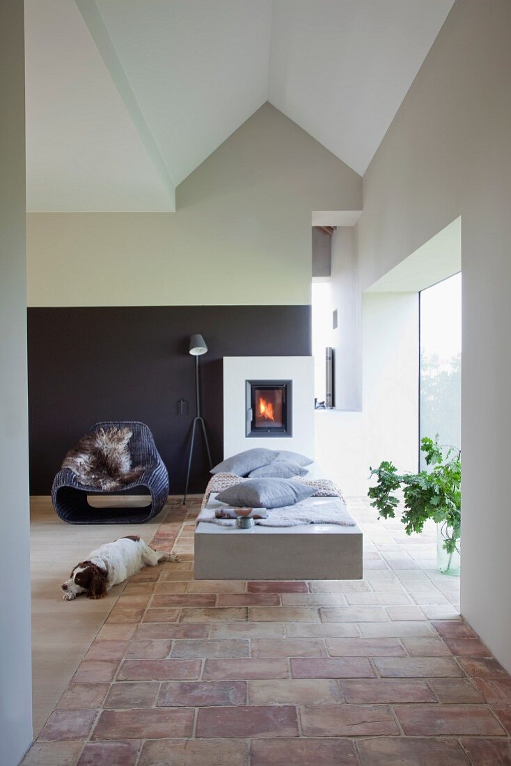 Dog lying next to concrete chaise on terracotta floor tiles in front of fireplace in modern interior