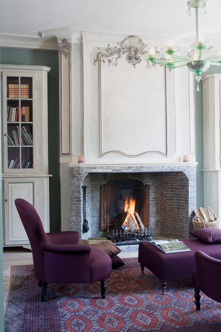 Elegant armchair and ottoman with purple upholstery in front of fireplace in grand interior
