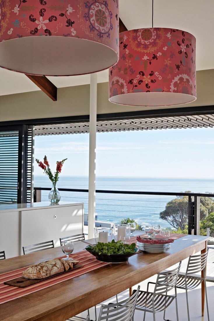 Pendelleuchten mit gemustertem Stoffschirm über Esstisch mit Metallstühlen; Panoramafenster mit Meerblick
