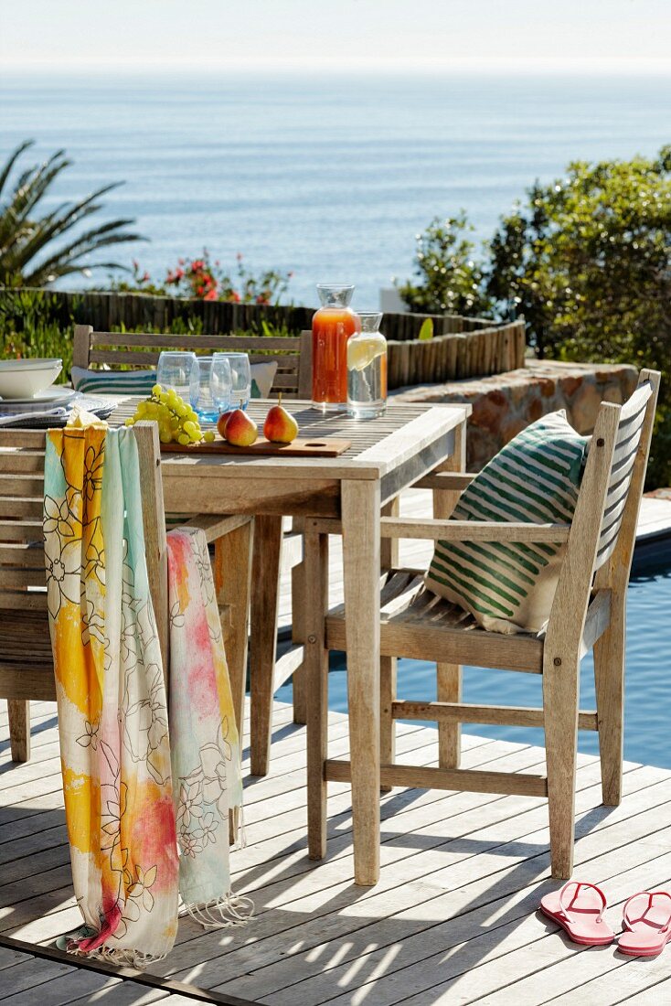 SonnenbeschieneneTerrasse mit Tisch und Stühlen aus Holz neben Pool, im Hintergrund das Meer