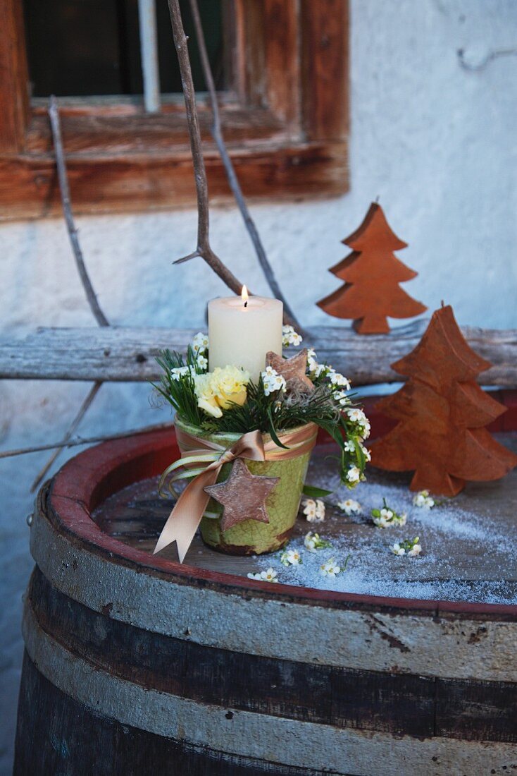 Kerzengesteck mit Blüten und Schleifenband in Übertopf auf Vintage-Fass mit Deko-Holzbäumchen