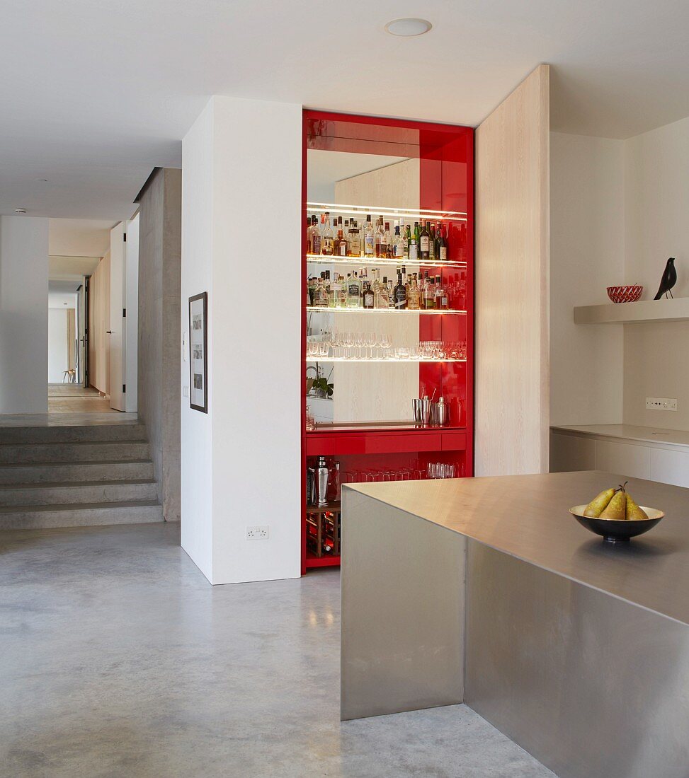 View across stainless steel kitchen counter to floor-to-ceiling home bar with swivelling door and red interior and into corridor with concrete steps
