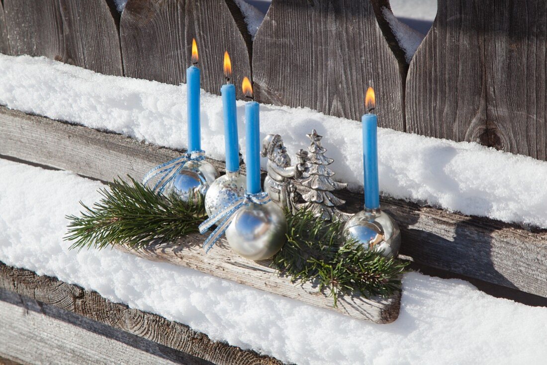 Adventsgesteck mit glänzenden Weihnachtskugeln als Kerzenhalter auf verschneiter, verwitterter Holzunterlage