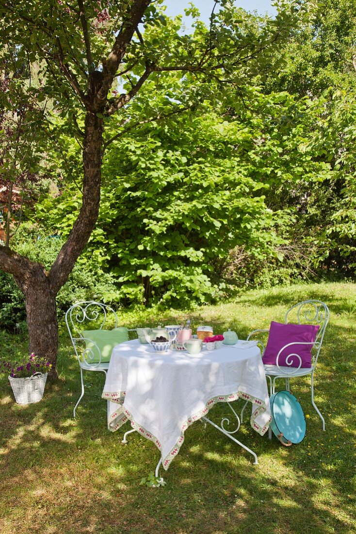 Filigrane Metallstühle um gedeckten Tisch mit Tischdeke und Häkelborte unter schattigem Baum