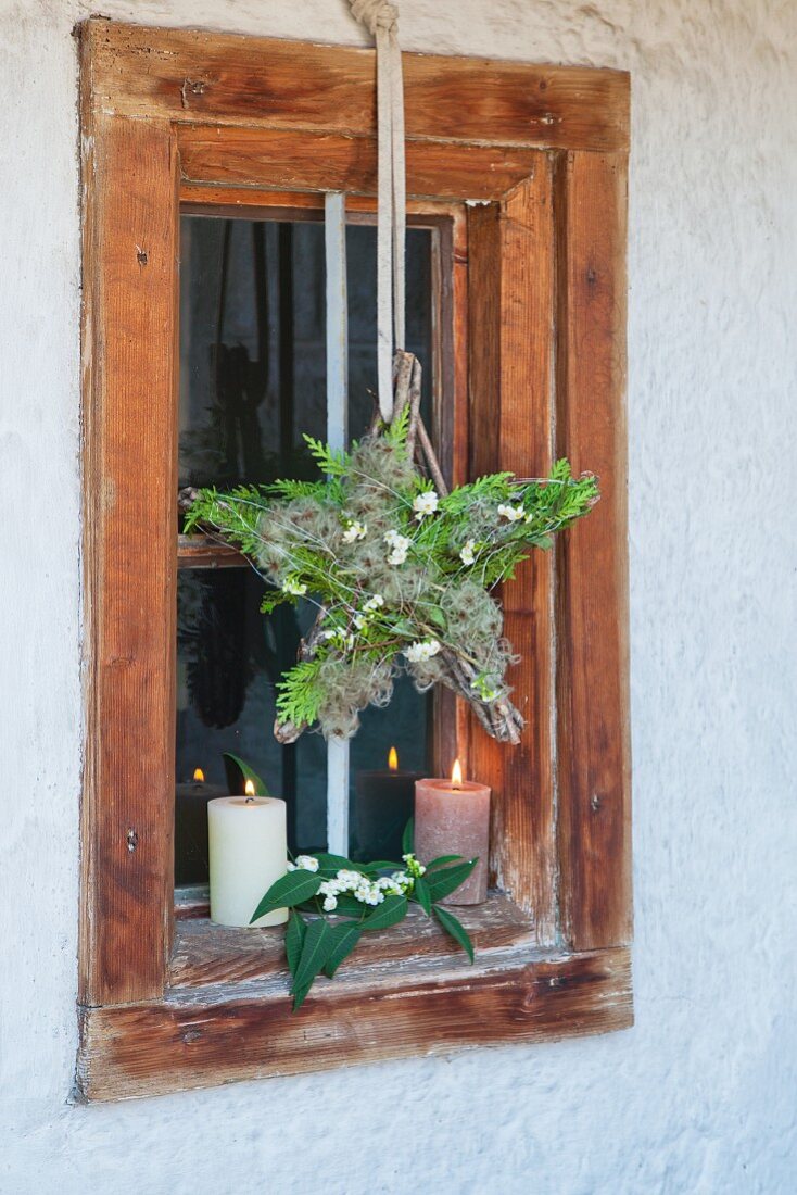 Aufgehängter, weihnachtlicher Fensterstern mit Koniferengrün und Clematisfruchtständen an rustikalem Fenster mit Kerzenlicht