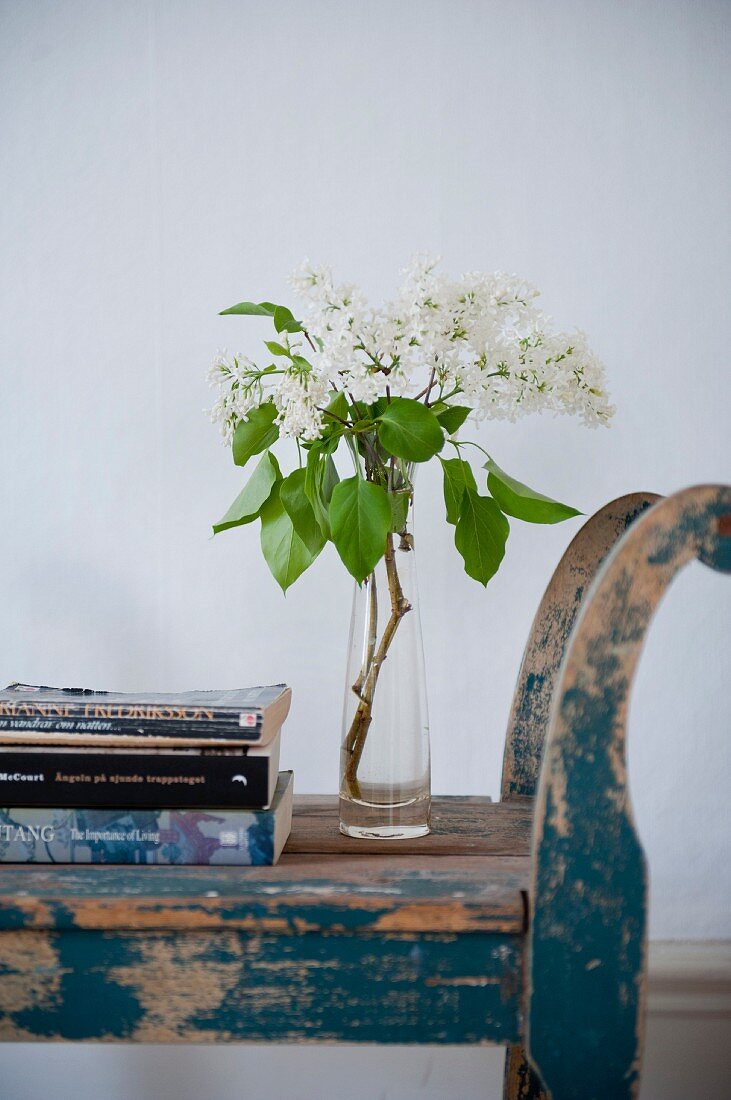 Glass vase of white lilac and stack of books on vintage blue wooden bench