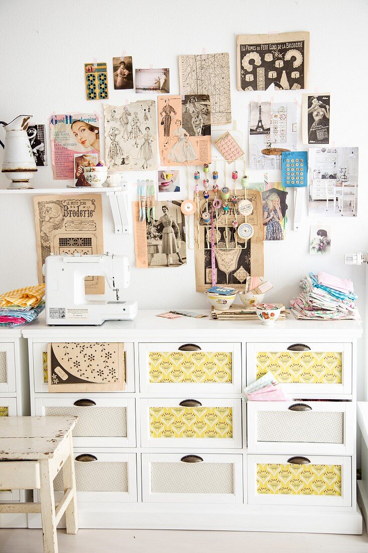 Wall decorated with clipping from retro fashion magazines above sewing utensils and sewing machine on top of white chest of drawers