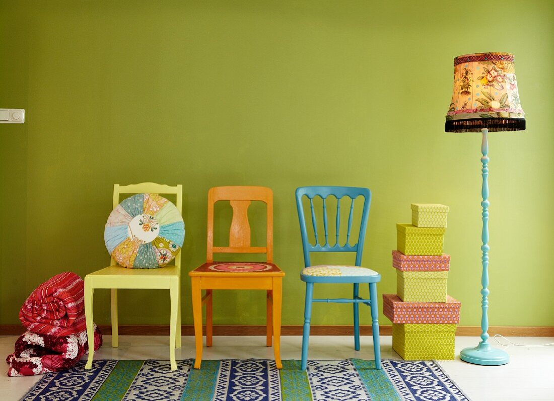Bunte Holzstühle mit Patchworkkissen, Schachtelstapel und Vintage-Stehleuchte vor hellgrüner Wand