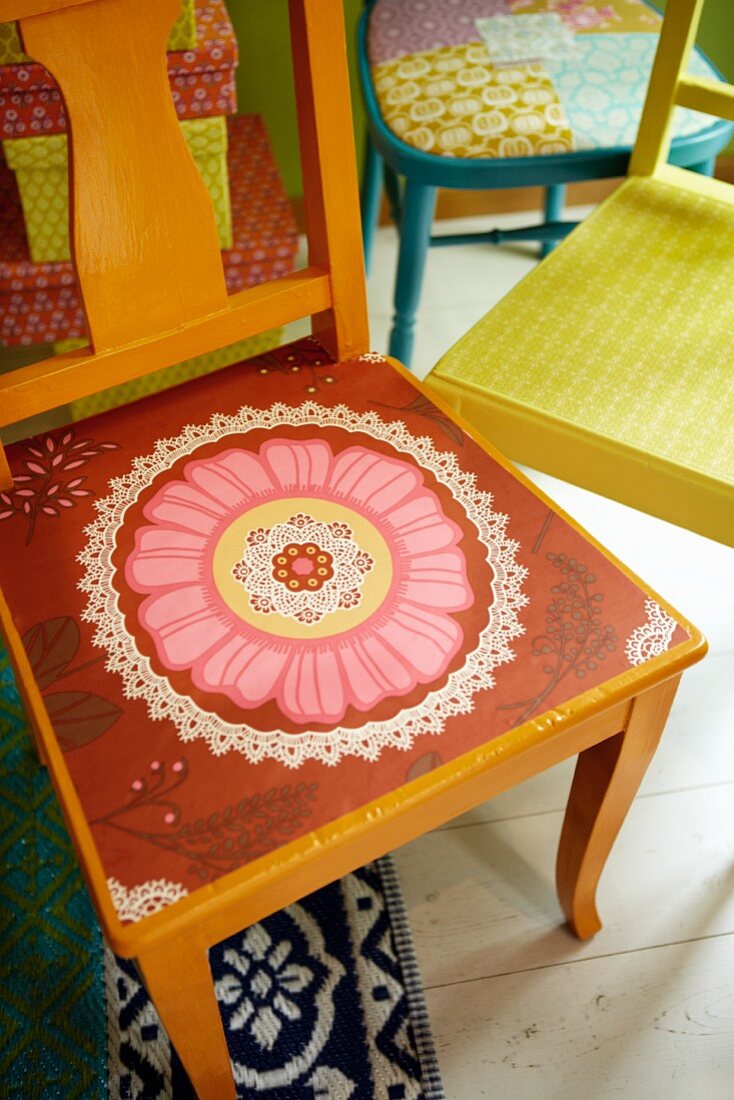Seat of orange-painted wooden chair painted with pink and red floral motif in front of further colourful chairs