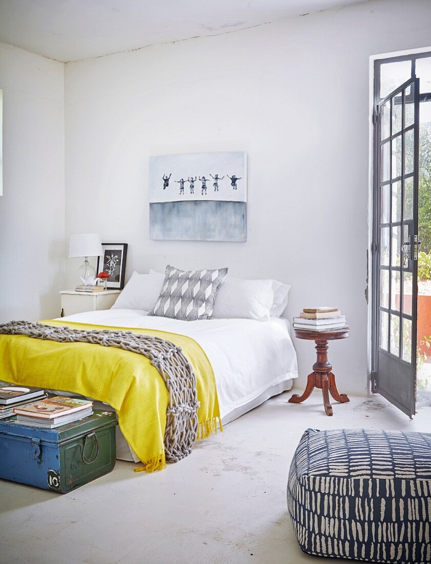 Schlafzimmer mit Doppelbett und gelber Tagesdecke, antikem Nachttisch und Sitzpouf mit gemustertem Bezug