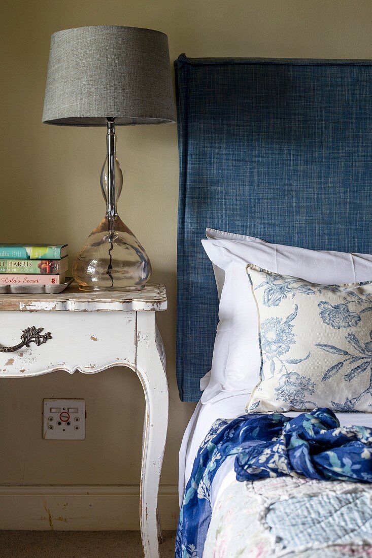 Table lamp with grey lampshade on vintage bedside table next to bed with headboard upholstered in blue fabric