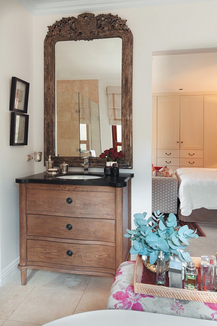 Wooden chest of drawers with integrated sink below mirror with carved wooden frame next to floor-to-ceiling doorway with view into bedroom