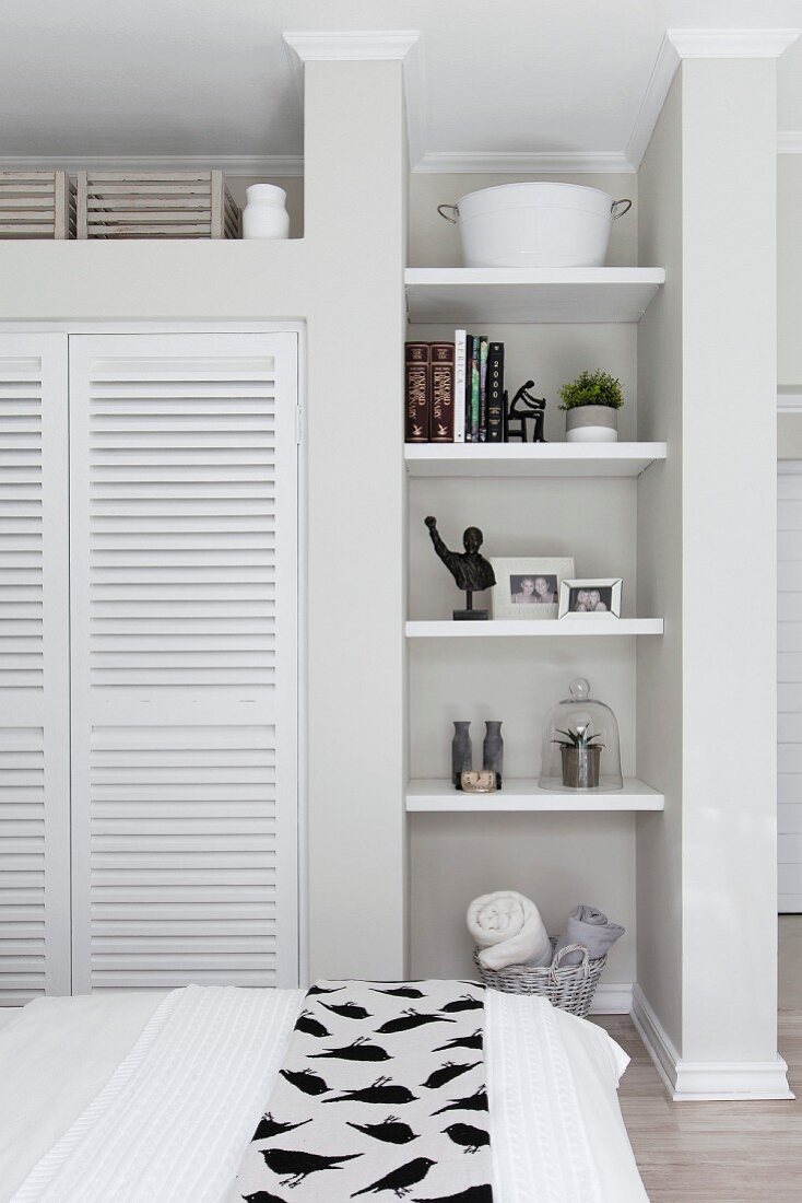 View over bed of shelves in narrow niche and fitted wardrobe with white louvre doors