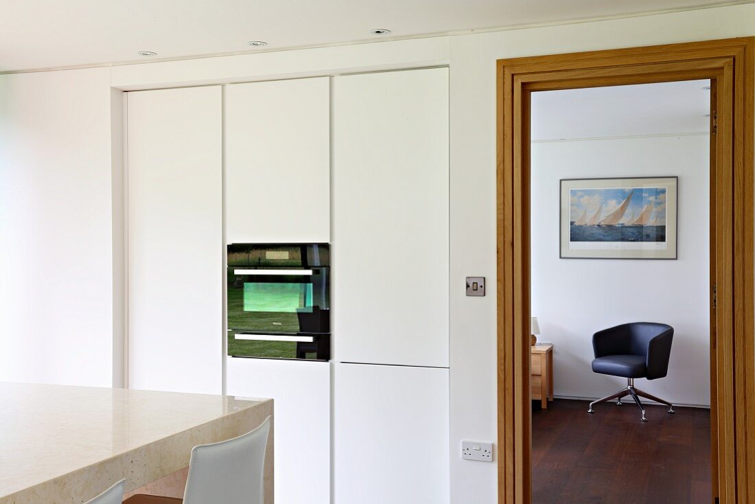 Weißer Kücheneinbauschrank mit Küchengerät neben offener Tür und Blick auf schwarzen Drehsessel