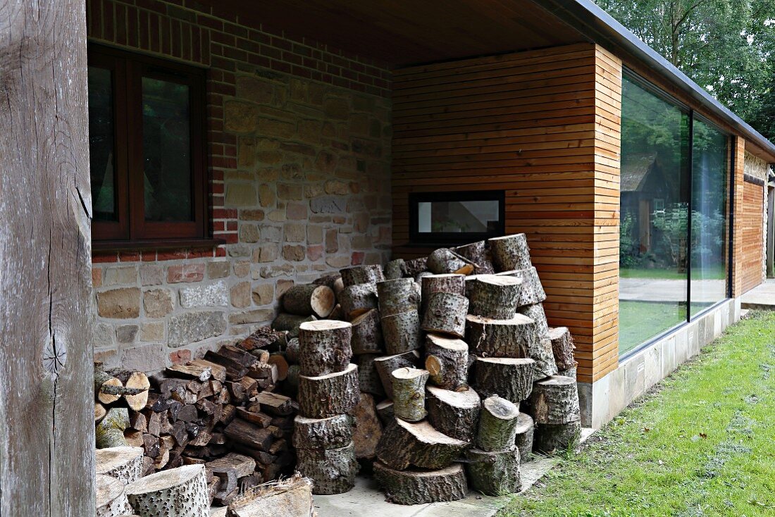 Firewood stacked in façade niche of modern house with wooden cladding and stone walls