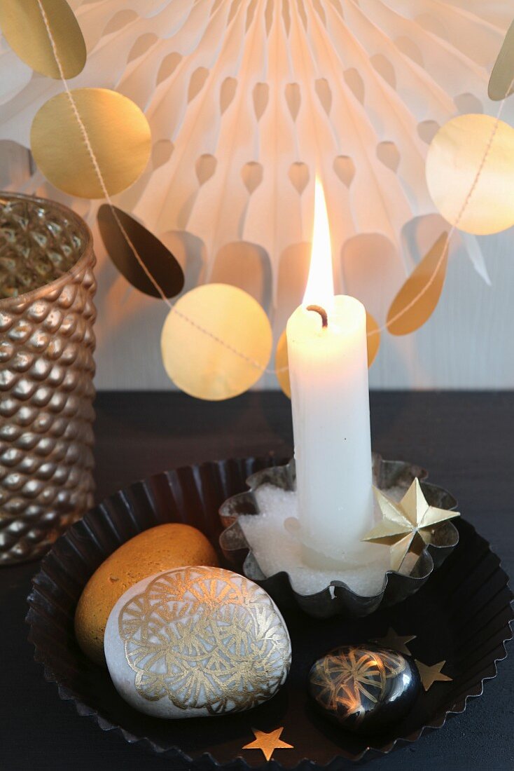 Lit candle and gold-painted pebbles in black cake tin in front of garland of gold paper discs