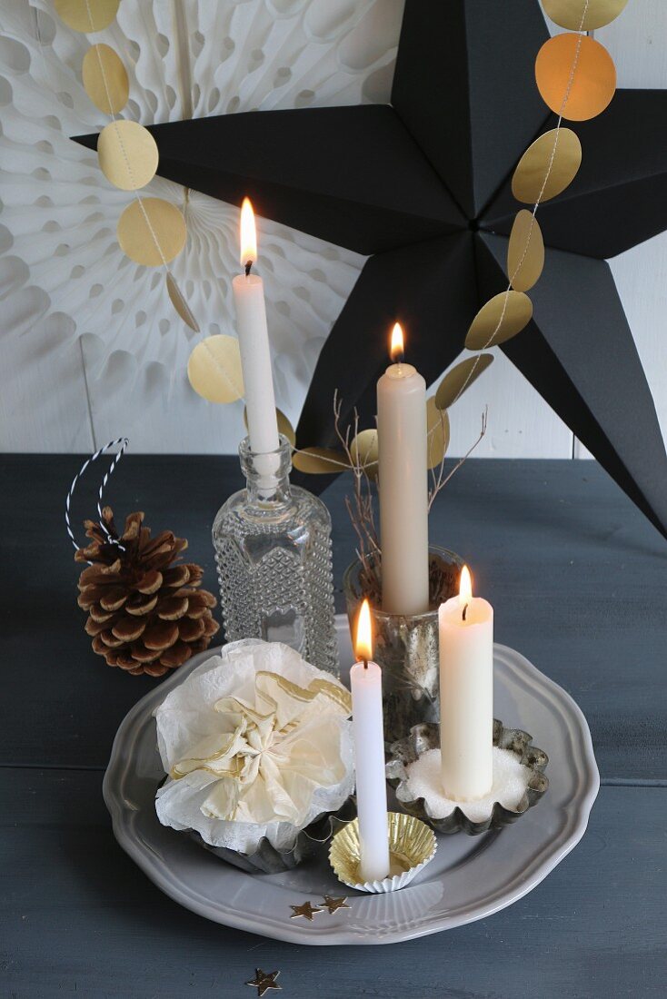 Weihnachtsdekoration und Adventskranzidee, vier brennende Kerzen in verschiedenen Gefässen um Papierblume, vor Girlande aus goldenen Papierplättchen